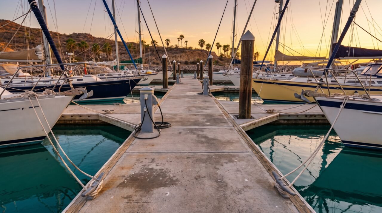 Muelle flotante de una marina al atardecer con veleros amarrados a ambos lados, cornamusas y pedestales de electricidad en primer plano