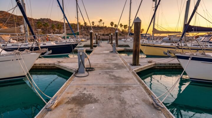 Muelle flotante de una marina al atardecer con veleros amarrados a ambos lados, cornamusas y pedestales de electricidad en primer plano