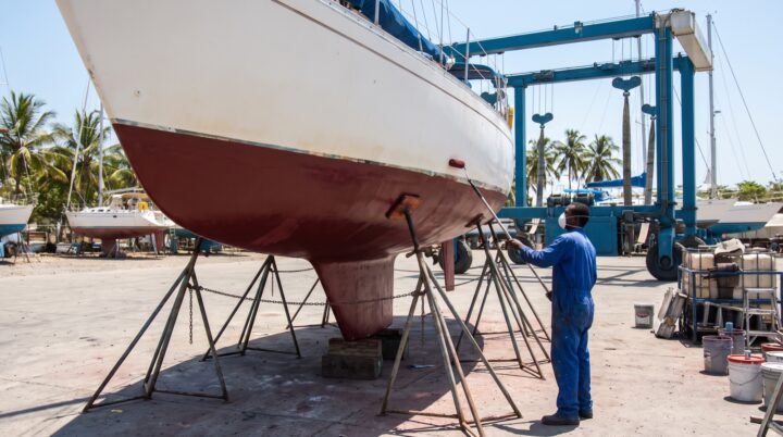 Velero de crucero en calzos en un varadero tropical mientras un operario aplica antifouling al casco, con un travel lift al fondo
